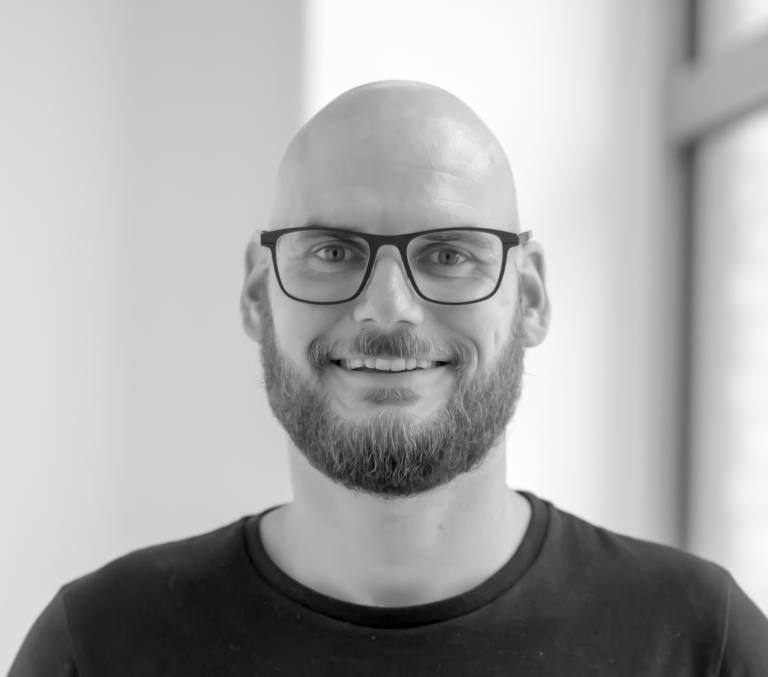 Porträt Christian Wieloch Mann mit Glatze und Brille, lächelt vor einer hellen Wand.